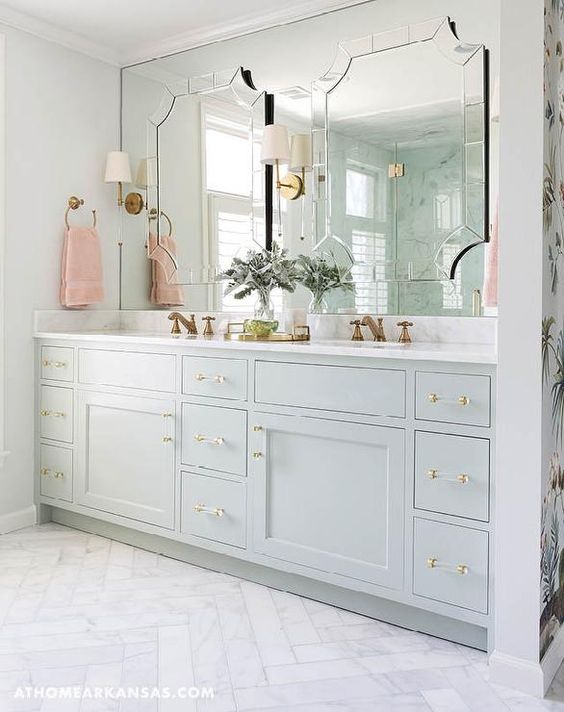 A white marble herringbone tile floor leads to a stunning bathroom boasts a pale gray dual washstand painted Sherwin Williams Contented adorned with brass and glass pulls topped with Alabama White Marble fitted with his and hers sinks and aged brass faucets placed under a full length mirror accented with individual mirrors illuminated by Camille Long Sconces.