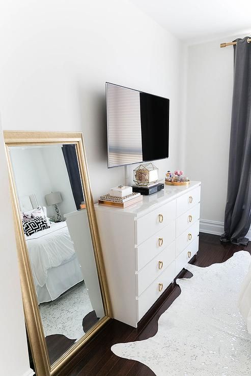 Chic bedroom features a flatscreen TV atop a white Ikea Malm Dresser adorned with gold ring hardware next to a gold leaf leaning mirror alongside a metallic cowhide rug.