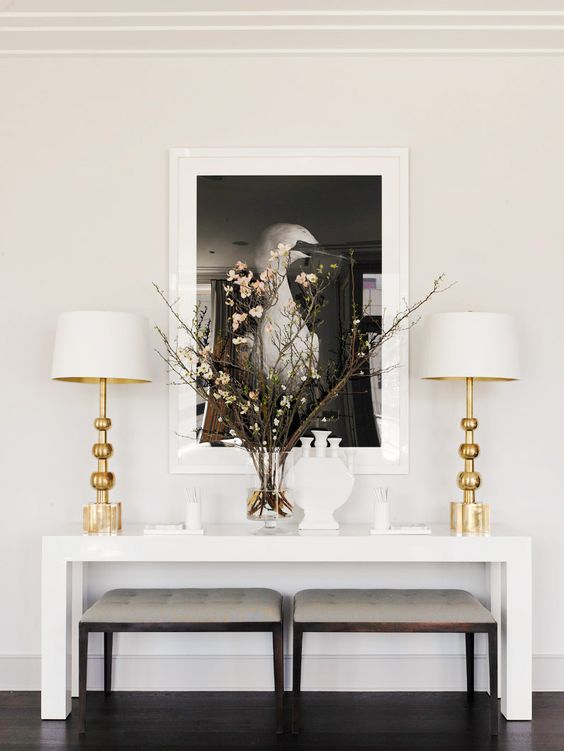 White and gold lamps on top of a white table with a glass vase and black and white wall art