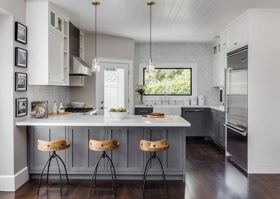White and gray kitchen features gray distressed cabinets paired with white marble countertops and a white and gray marble herringbone tile backsplash. A farmhouse sink and deck mount faucet stands under a picture window next to a corner glass door which leads to the backyard. A shiplap ceiling is accented with two glass pendants which hang over a gray kitchen peninsula lined with Arteriors Henson Counter Stools.