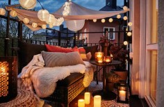 A cozy balcony with lit LED light chains, a canopy, lanterns with lit candles and a sofa filled with cushions and sheepskin rugs.