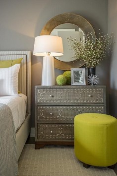 Contemporary yellow and gray bedroom features a gray wall lined with a gray pins tripe headboard with nailhead trim on bed dressed in gray chevron blankets and yellow pillows beside a Gray Raffia Chest doubling as nightstand topped with Regina Andrew White Folded Glass Lamp under a round gold mirror alongside a round canary yellow stool. 