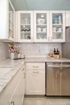 This light and bright modern kitchen combines white and gray tones to create a lively area of the home. Glass front cabinets are mixed in with white Shaker style cabinets, while a gray and white marble countertop offers a finishing touch. A white tile herringbone backsplash adds fun texture to the space.