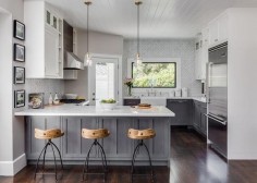 White and gray kitchen features gray distressed cabinets paired with white marble countertops and a white and gray marble herringbone tile backsplash. A farmhouse sink and deck mount faucet stands under a picture window next to a corner glass door which leads to the backyard. A shiplap ceiling is accented with two glass pendants which hang over a gray kitchen peninsula lined with Arteriors Henson Counter Stools.