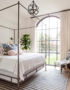 Lovely bedroom features a metal canopy bed, Oly Studio Marco Bed, dressed in soft white bedding and pink and gray ikat pillows as well as a pink ikat bolster pillow placed atop a beige and navy striped rug illuminated by glass vintage barn wall sconces and a Suzanne Kasler Morris Lantern.